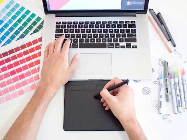 A designer working on a drawing tablet beside a laptop, with colour swatches on the desk